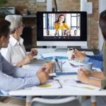 A group of colleagues sit around a table in an office, taking part in a video conference. They are watching and listening to a remote speaker displayed on a large screen during a work meeting.