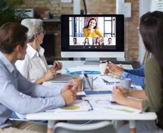 A group of colleagues sit around a table in an office, taking part in a video conference. They are watching and listening to a remote speaker displayed on a large screen during a work meeting.
